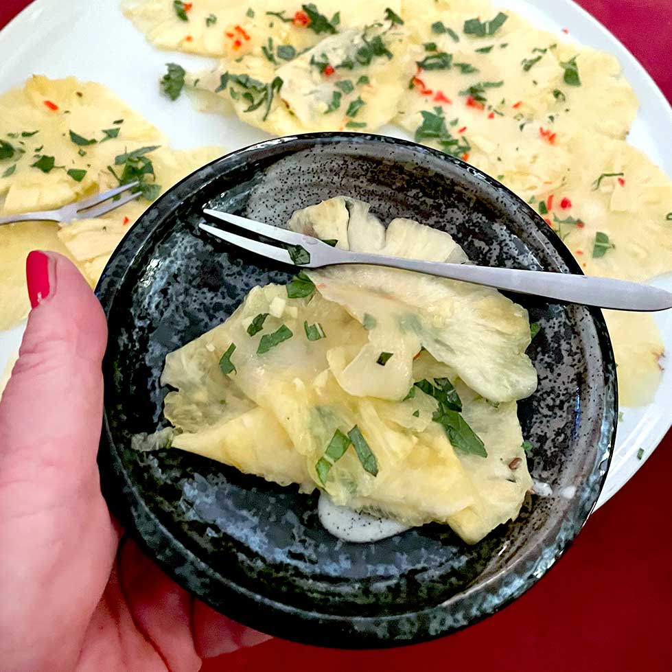 A hand holds a dark ceramic bowl with thinly sliced pineapple, garnished with fresh herbs. In the background, more pineapple slices with herbs and red chilli are arranged on a white plate. A small fork rests in the bowl.