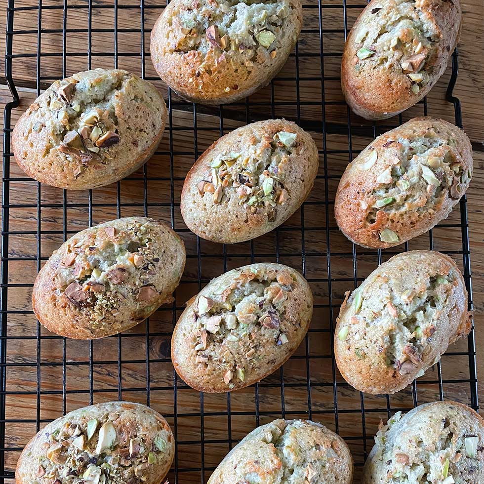 Oval-shaped baked goods topped with chopped nuts are cooling on a black wire rack placed on a wooden surface. The golden-brown pastries appear fluffy and freshly baked.