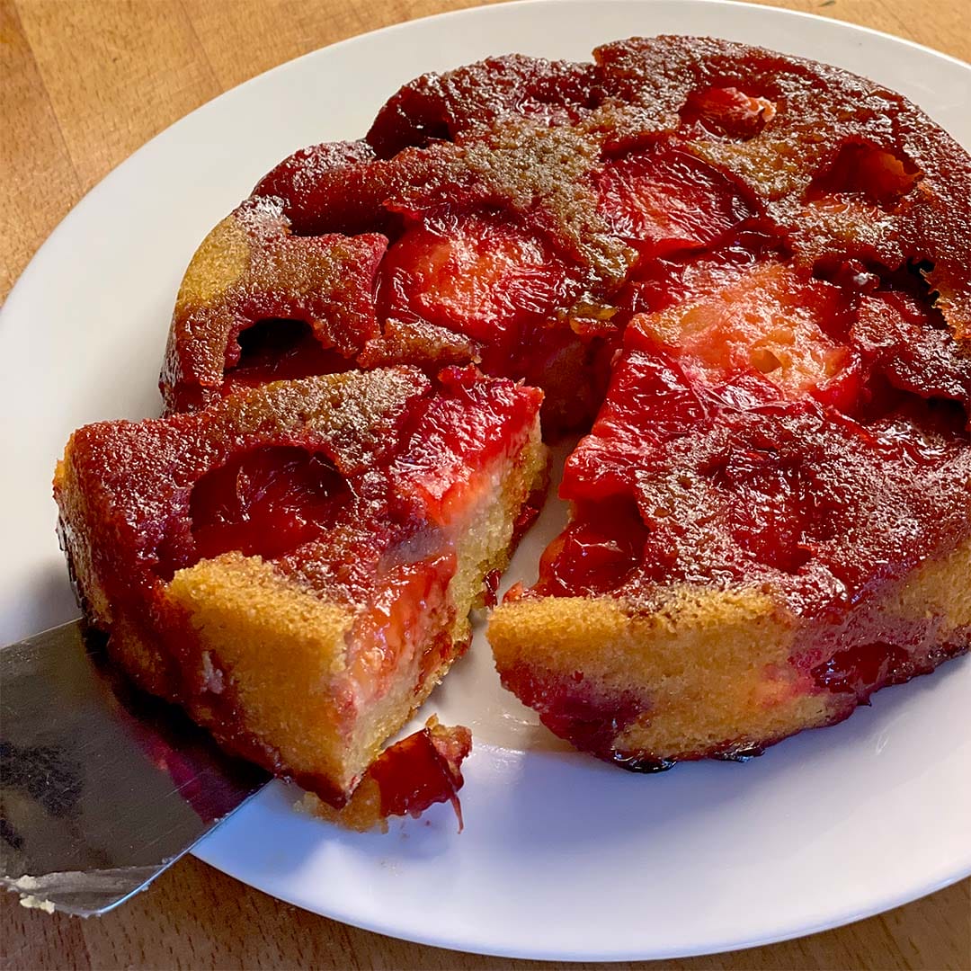 A close-up of a plum upside-down cake on a white plate, with a slice being lifted out. The cake has caramelised plums on top and a golden, moist-looking base. The background is a wooden surface.