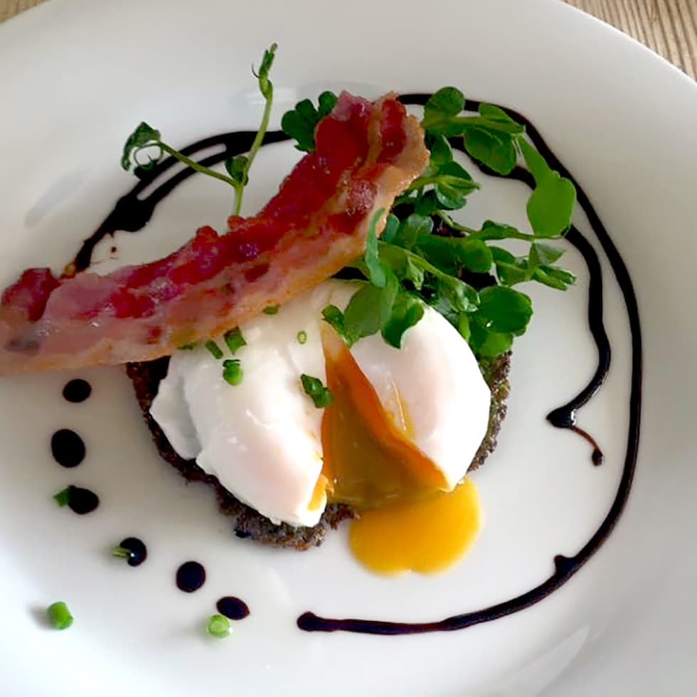 A poached egg with a runny yolk sits on a dark patty, garnished with microgreens and chives, topped with a crispy rasher of bacon, and surrounded by a swirl of dark sauce on a white plate.