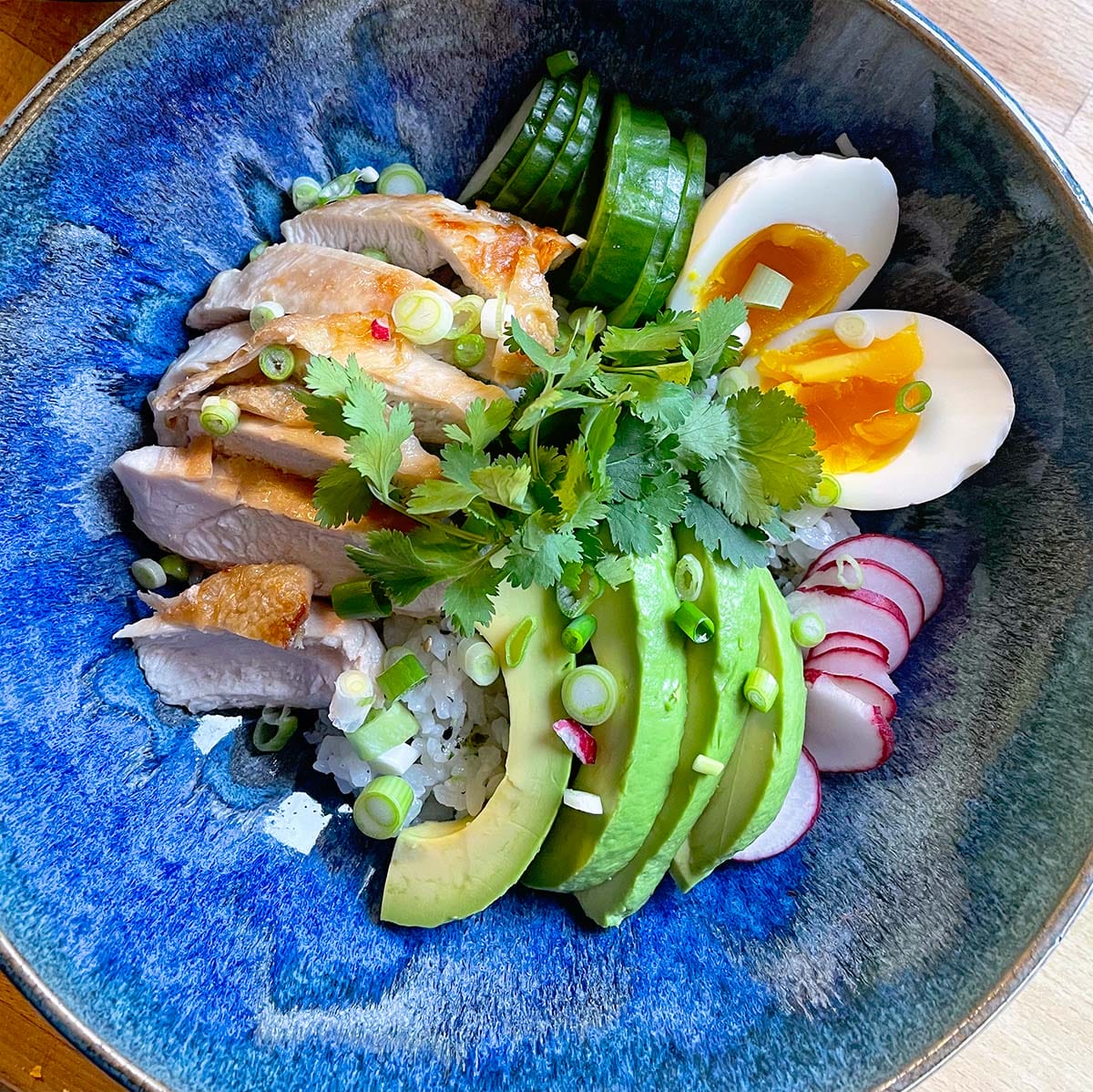 A blue bowl filled with rice, sliced grilled chicken, avocado, soft-boiled egg, cucumber, radish, coriander, and spring onions, arranged neatly for a colourful and fresh meal.