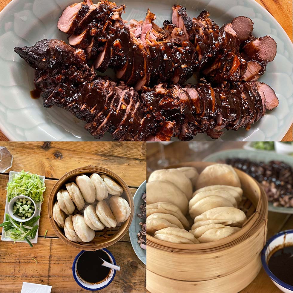 A platter of sliced, glazed pork fillet, a bamboo steamer filled with fluffy bao buns, and plates of spring onions, cucumber, and hoisin sauce arranged on a wooden table.