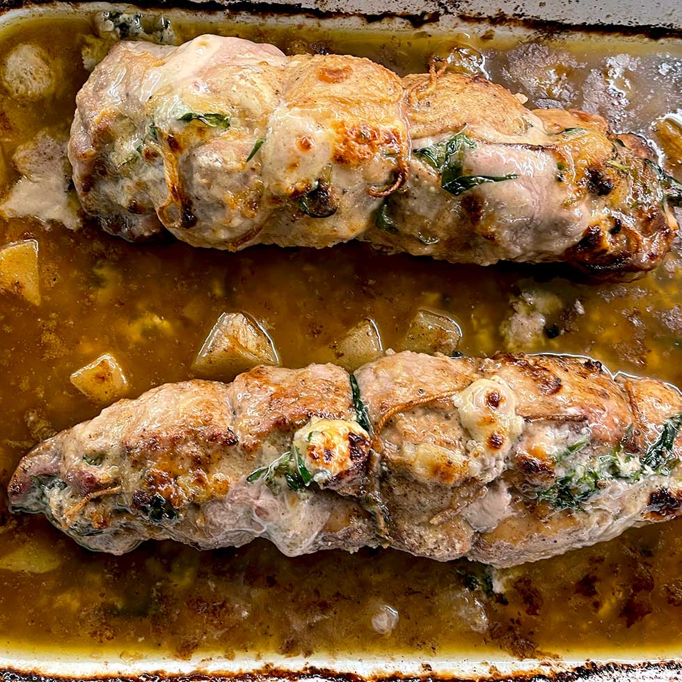 Two stuffed pork fillets tied with string are baked in a brown sauce with visible herbs and chunks of vegetables in a rectangular roasting tin.