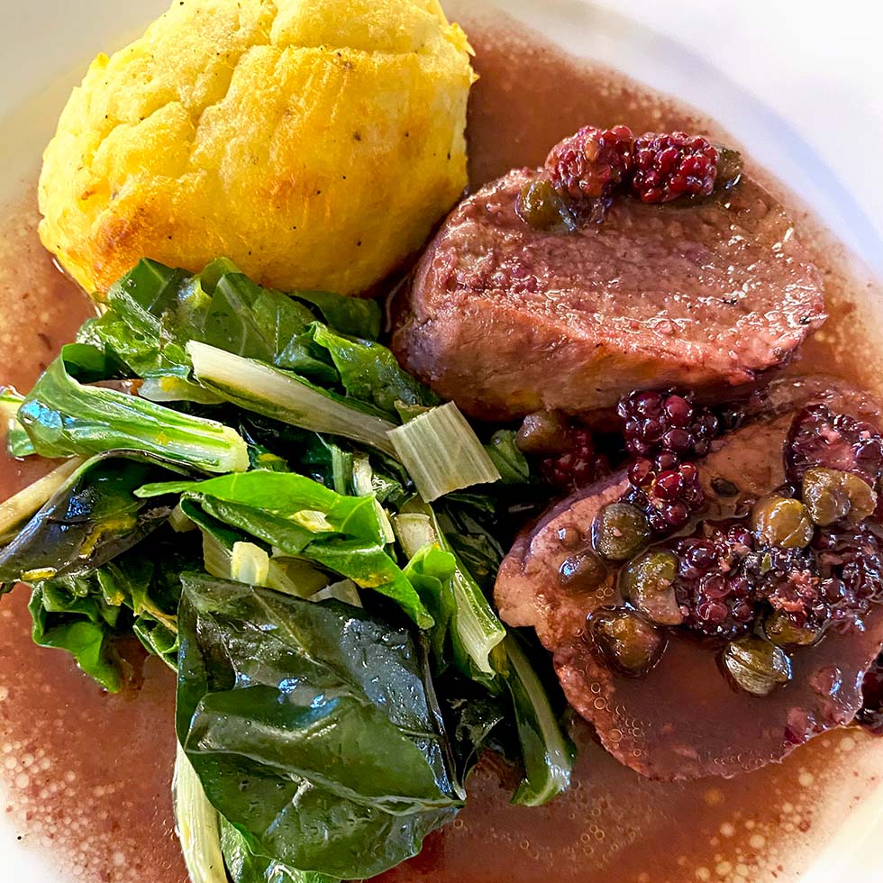 A plate with two slices of beef topped with capers and berries in sauce, sautéed leafy greens, and a round serving of cornbread or a baked yellow side.