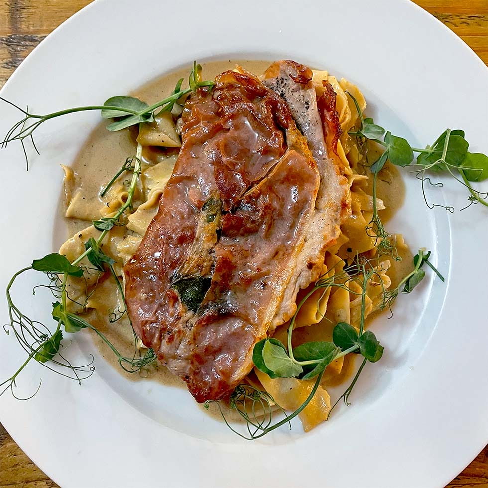 A white plate with pasta topped with a slice of prosciutto-wrapped meat, creamy sauce, and garnished with fresh pea shoots.