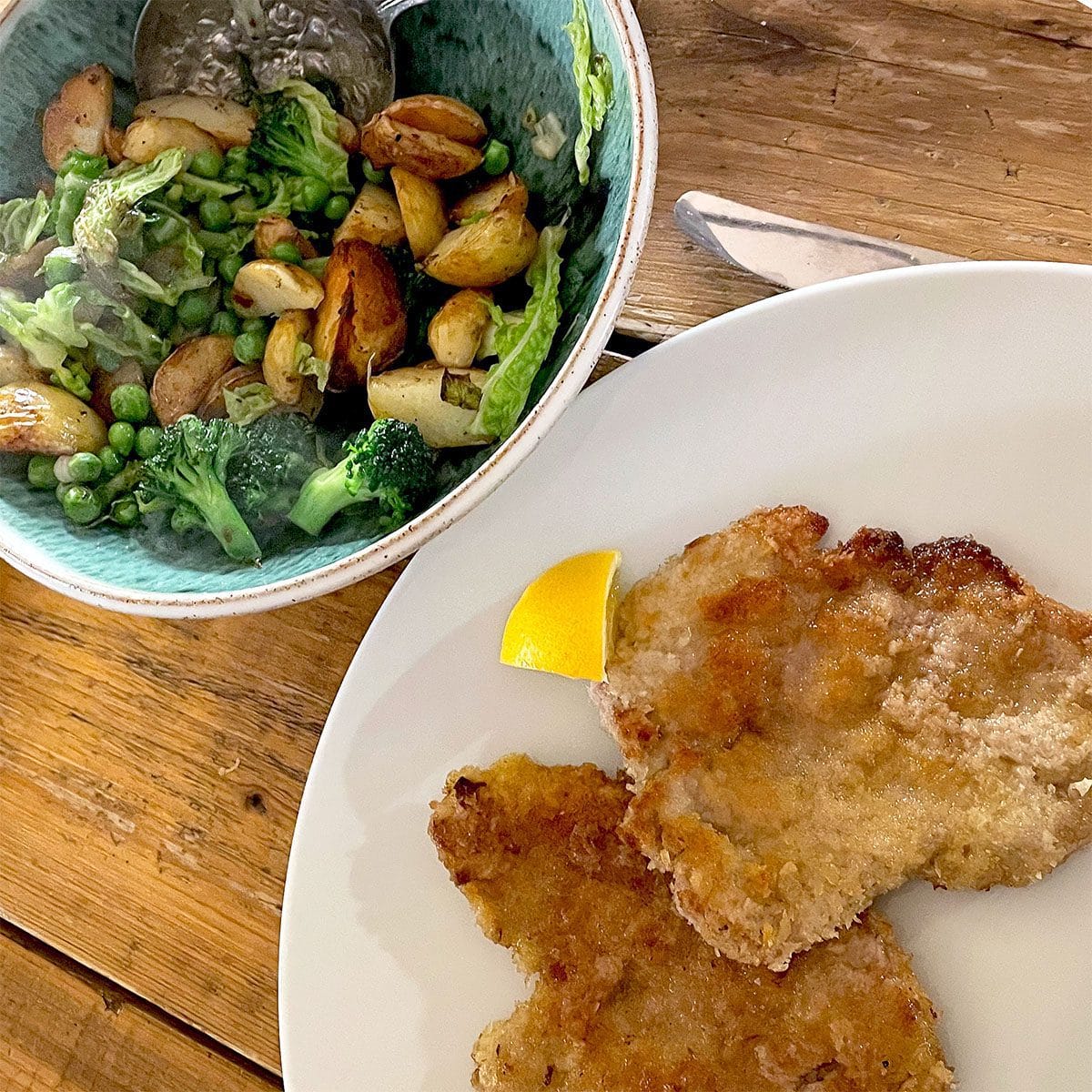 A white plate with two breaded cutlets and a lemon wedge, beside a bowl of roast potatoes, broccoli, peas, and greens on a wooden table.