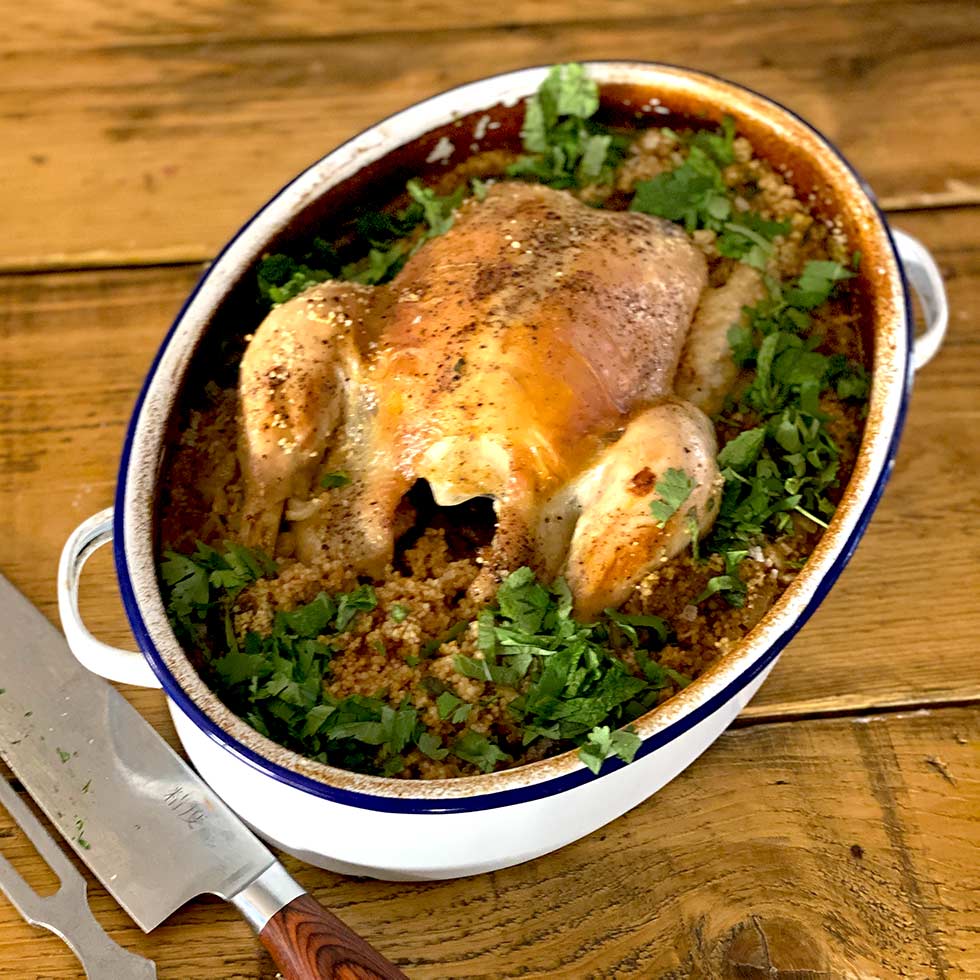 A whole roast chicken sits atop a bed of seasoned grains and herbs in an oval casserole dish, garnished with chopped parsley, on a wooden table beside a carving knife.