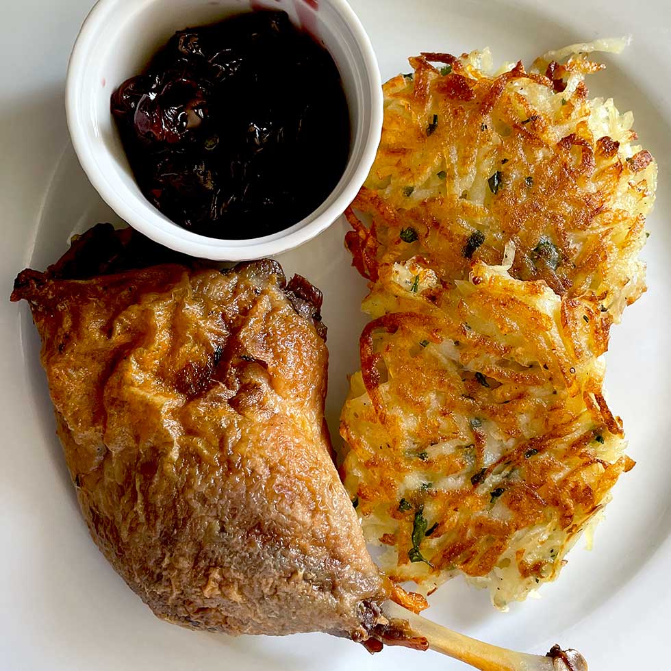 A plate with a crispy duck leg, two golden-brown potato latkes, and a small cup of dark fruit sauce, arranged on a white dish.