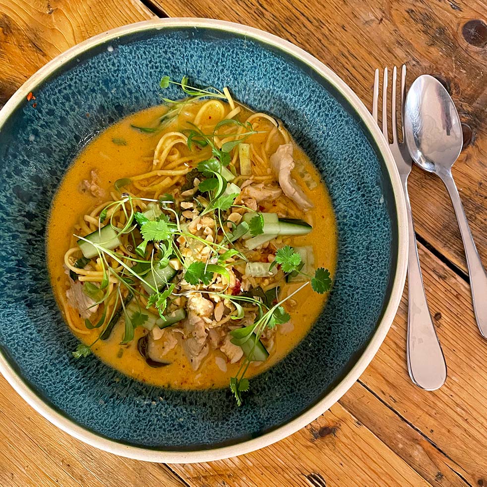 A bowl of noodle soup with sliced vegetables, herbs, and chicken in a creamy, orange broth, served on a rustic wooden table with a spoon and fork beside the bowl.