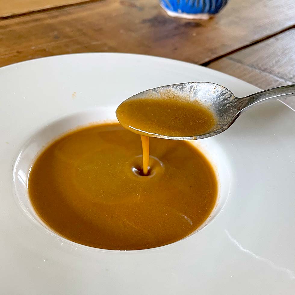 A spoon pours thick brown gravy into a shallow white bowl, with some gravy already pooled at the bottom. The scene is set on a wooden table with a blue and white cup in the background.
