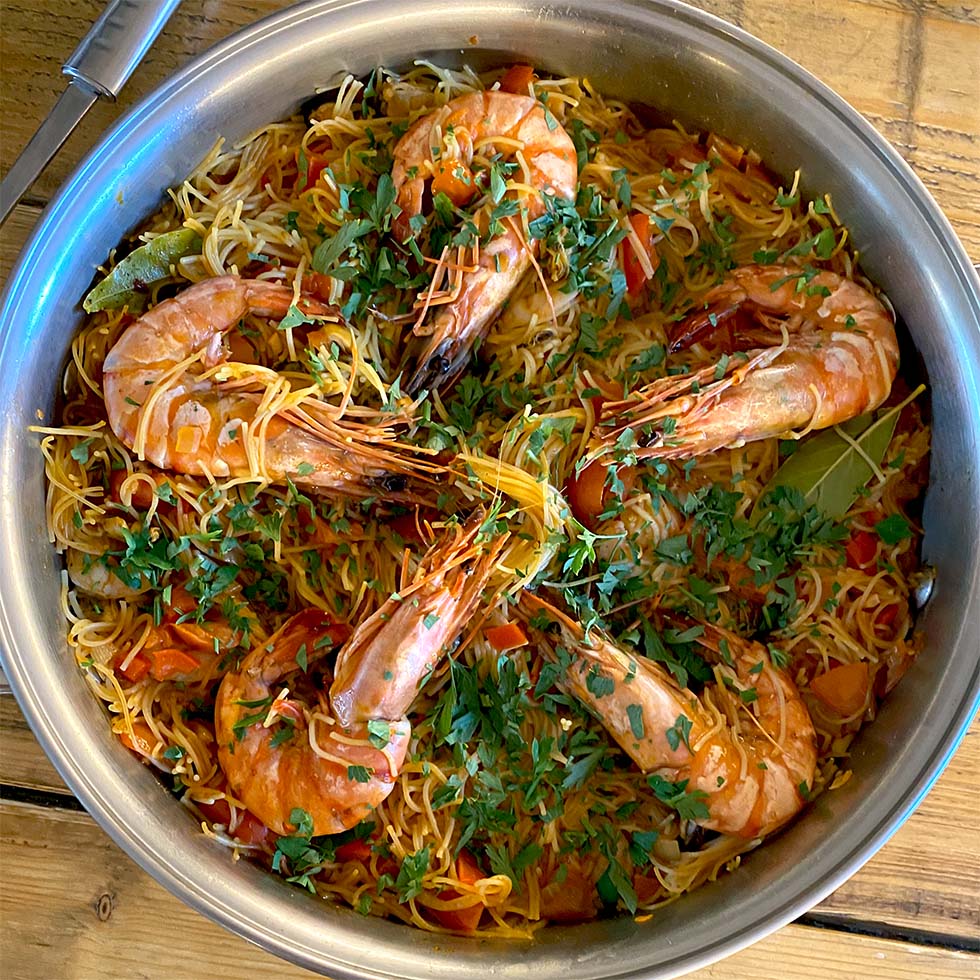 A metal pan filled with cooked noodles, large prawns, chopped tomatoes, and garnished with fresh herbs, sits on a wooden table.
