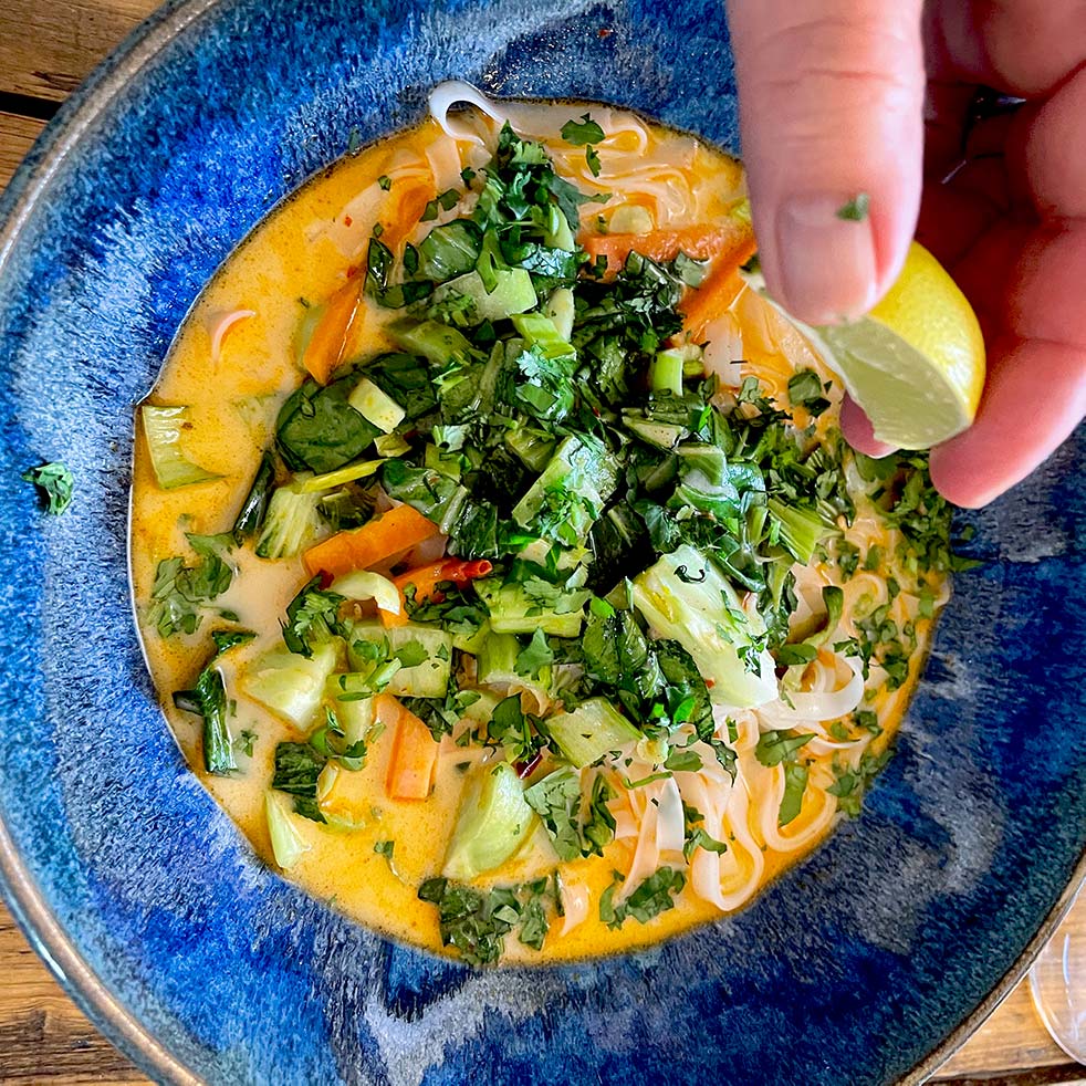 A hand squeezes a lime wedge over a bowl of colourful curry soup with noodles, leafy greens, carrots, and chopped fresh herbs in a blue dish.