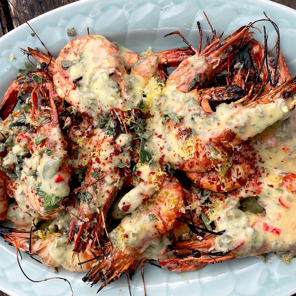 Grilled prawns with heads on, served on a white platter, topped with a creamy herb and lemon sauce, sprinkled with fresh herbs and red pepper flakes.