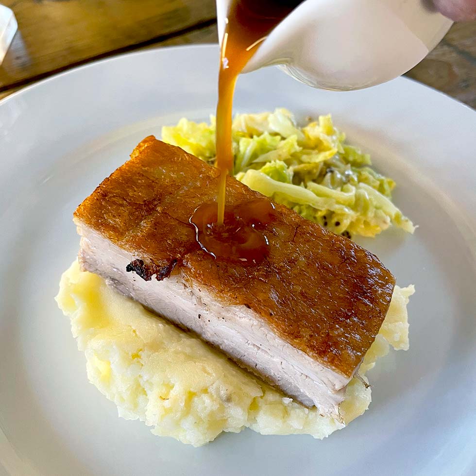 A thick slice of crispy roast pork belly sits on mashed potatoes, with gravy being poured over it. A side of cooked cabbage is also on the white plate.