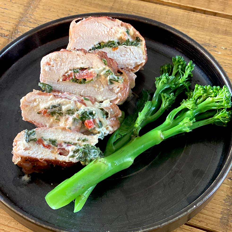 Sliced stuffed chicken breast filled with spinach, cheese, and red peppers, served on a dark plate with steamed tenderstem broccoli on the side, placed on a wooden table.