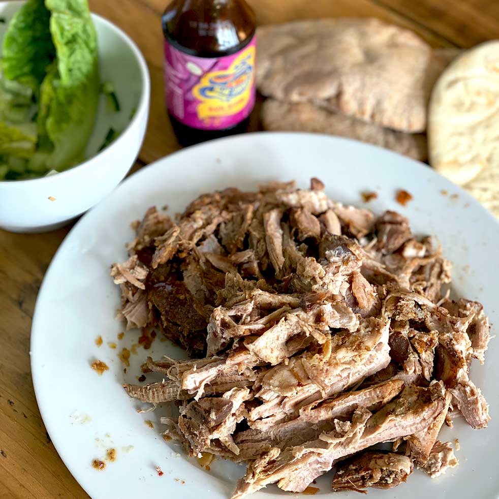 Shredded cooked meat on a white plate, with pitta bread, a bottle of sauce, and a bowl of lettuce and cucumber on a wooden table.