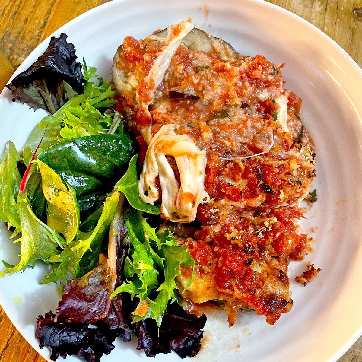 A white plate with a serving of aubergine Parmesan topped with tomato sauce and melted cheese, next to a side of mixed leafy green salad.
