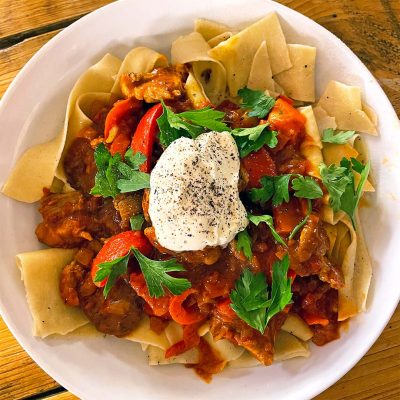 A white plate of wide pasta topped with a tomato-based meat stew, red peppers, fresh parsley, and a dollop of soured cream sprinkled with black pepper, served on a wooden table.