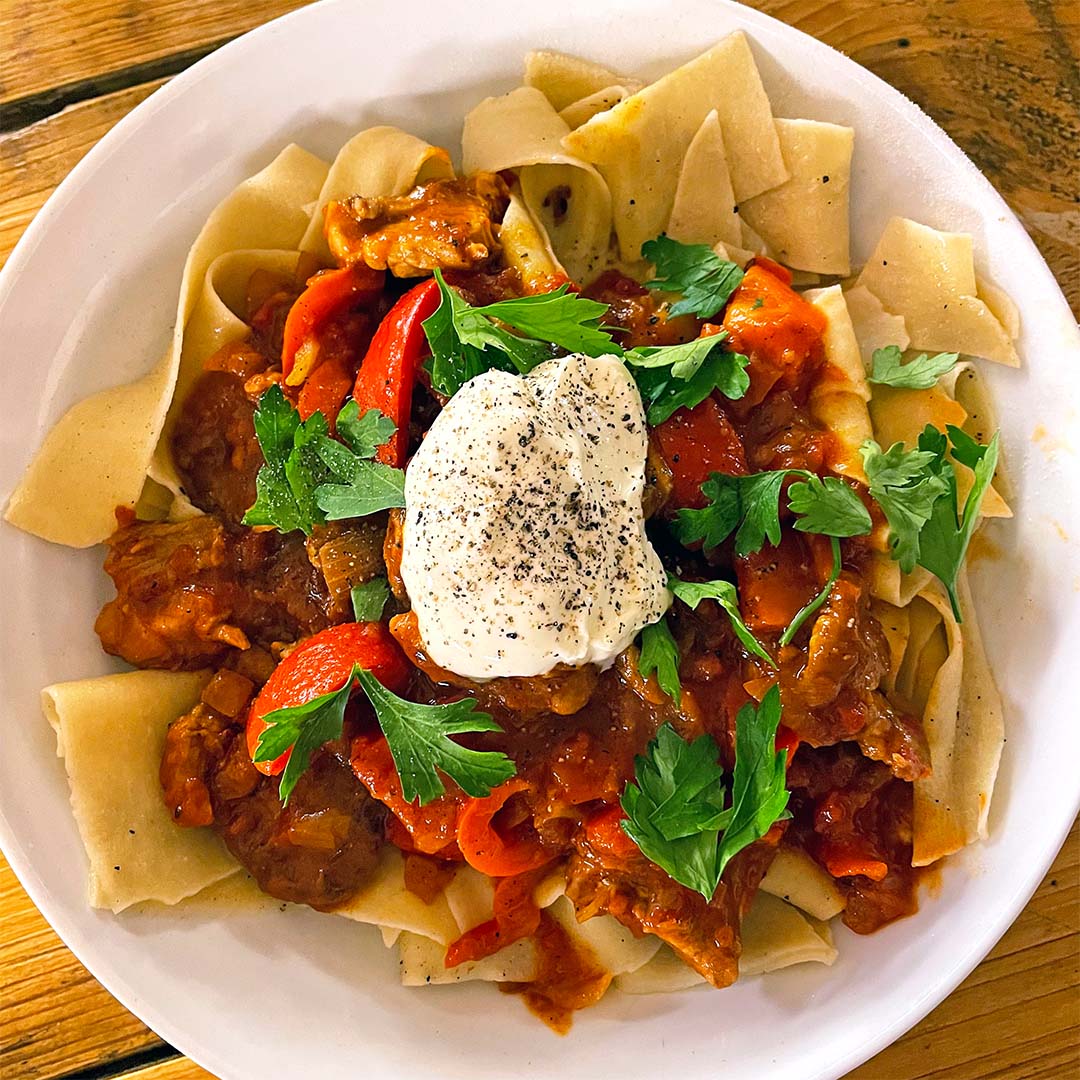 A white plate of wide pasta topped with a tomato-based meat stew, red peppers, fresh parsley, and a dollop of soured cream sprinkled with black pepper, served on a wooden table.