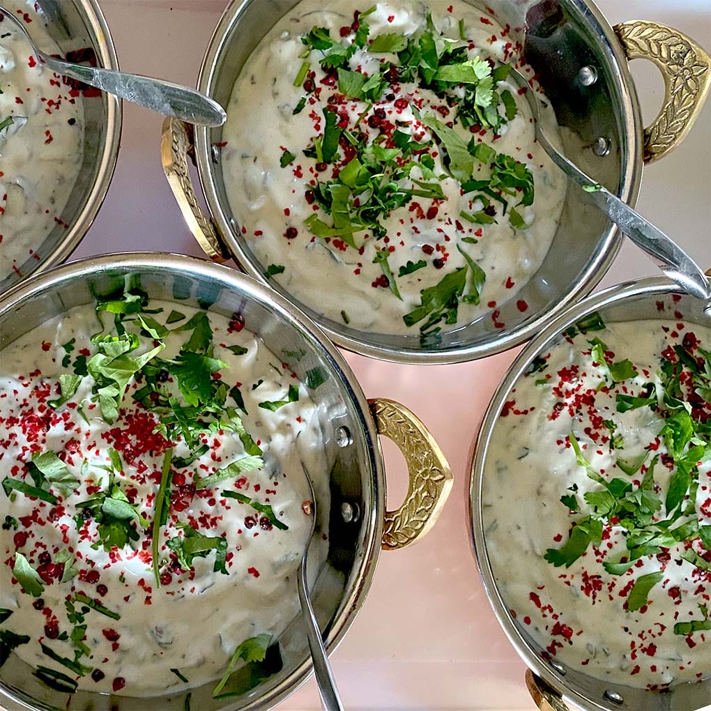 Four metal bowls with ornate handles are filled with creamy raita, garnished with chopped fresh herbs and red spices. Spoons rest in each bowl, and the bowls are arranged closely together on a light surface.