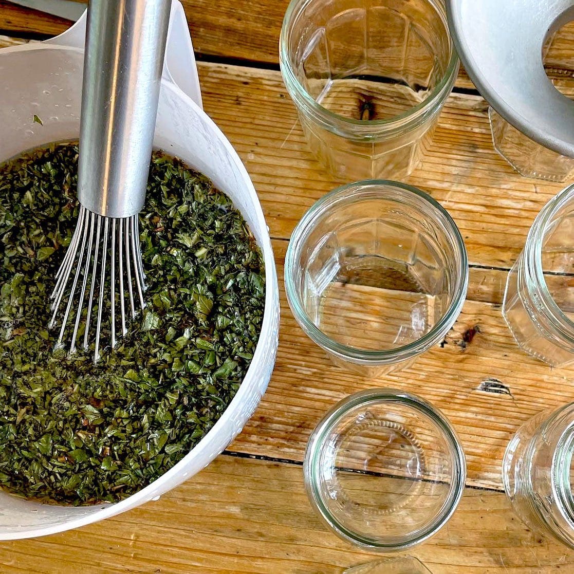 A metal whisk in a bowl of chopped green herbs sits on a wooden table next to several empty glass jars and a metal funnel, ready for filling.