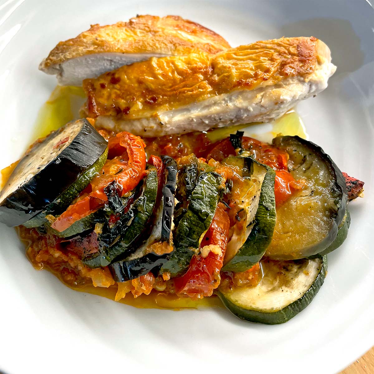 A plate with a sliced, golden-brown roasted chicken breast and a serving of colourful ratatouille made with courgette, aubergine, and tomatoes, all arranged neatly on a white dish.