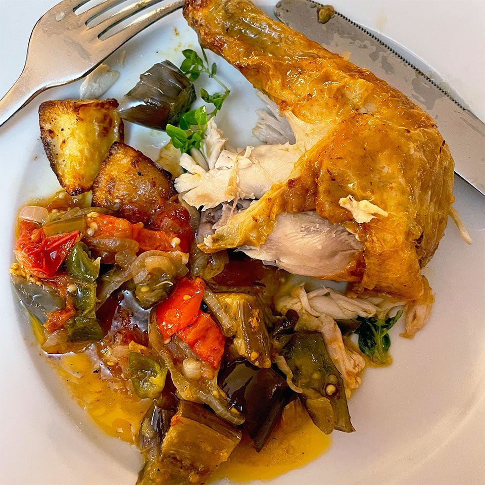 A plate with a roasted chicken leg, roast potatoes, and a colourful mix of cooked vegetables, including tomatoes, peppers, and aubergine, with a fork and knife on the side.