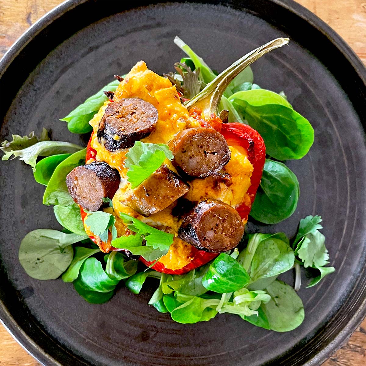 A halved red pepper filled with mashed potato and sliced sausage, garnished with fresh herbs and parsley, served on a dark plate with leafy greens underneath.
