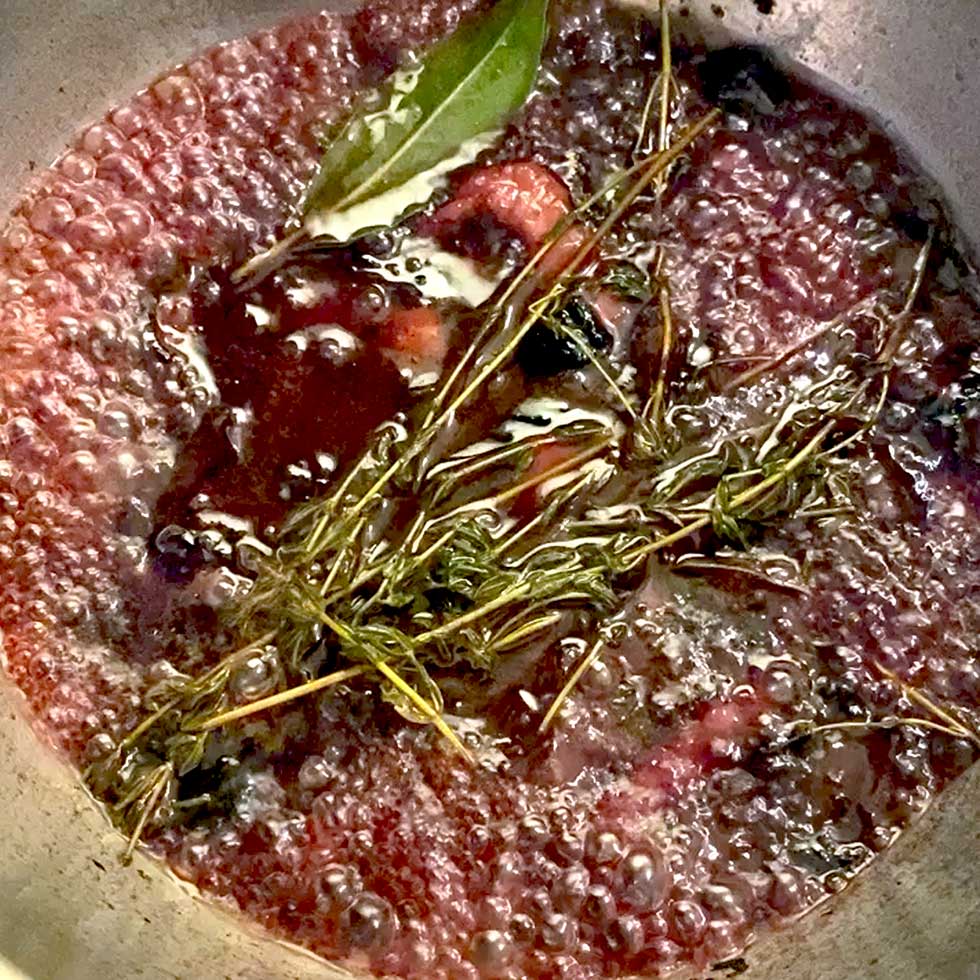 A close-up of a simmering pot with red wine, fresh thyme sprigs, a bay leaf, and chunks of vegetables, likely preparing a sauce or reduction. Bubbles are visible on the surface as it cooks.