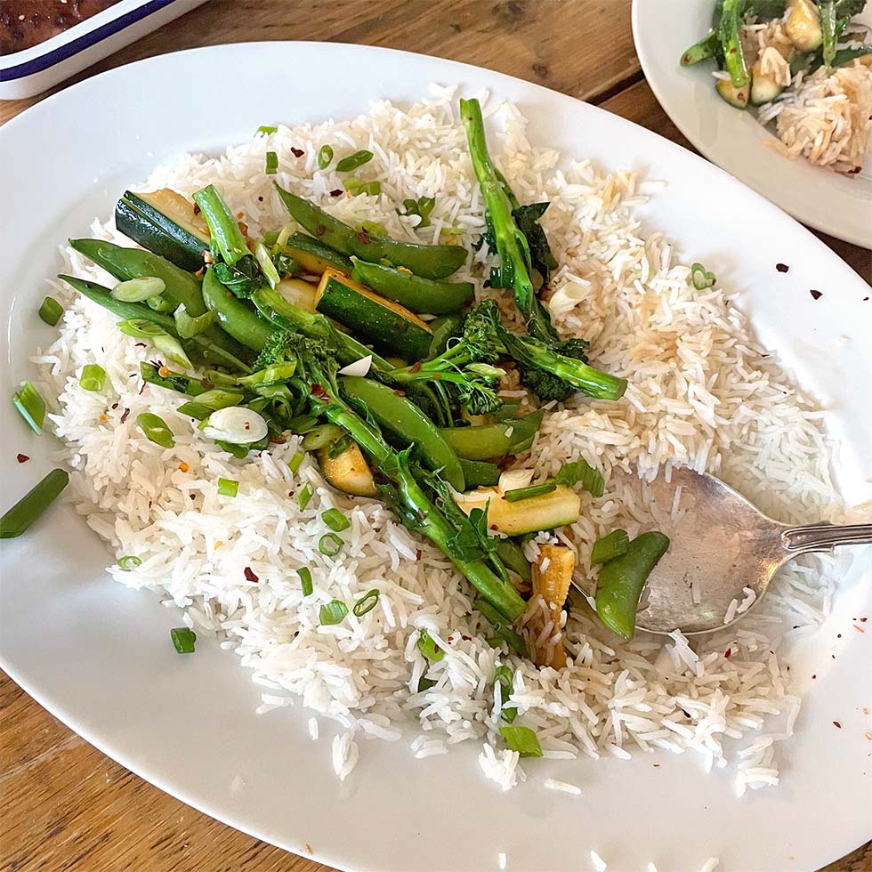 A white oval plate with steamed white rice topped with green vegetables, including sprouting broccoli, mangetout, and courgette, garnished with sliced spring onions and red chilli flakes. A serving spoon rests on the plate.