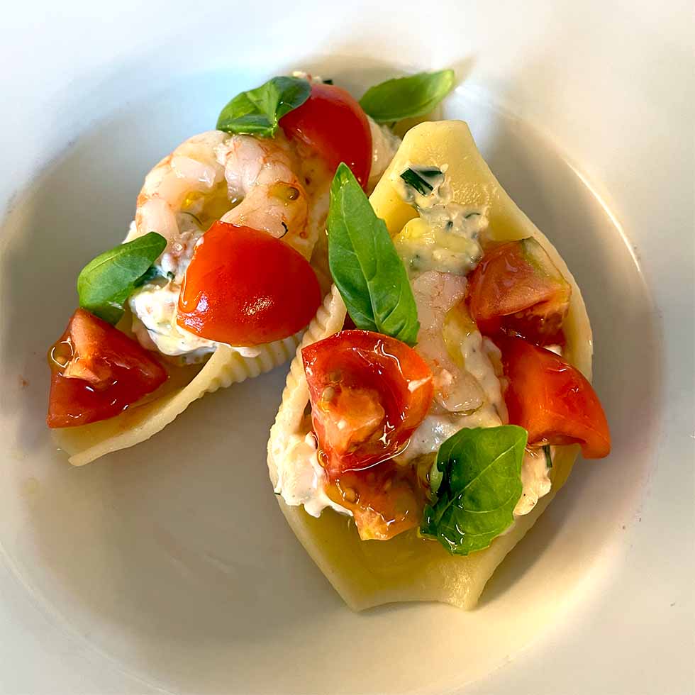 Two pasta shells filled with creamy cheese, prawns, chopped tomatoes, and fresh basil leaves, served on a white plate.