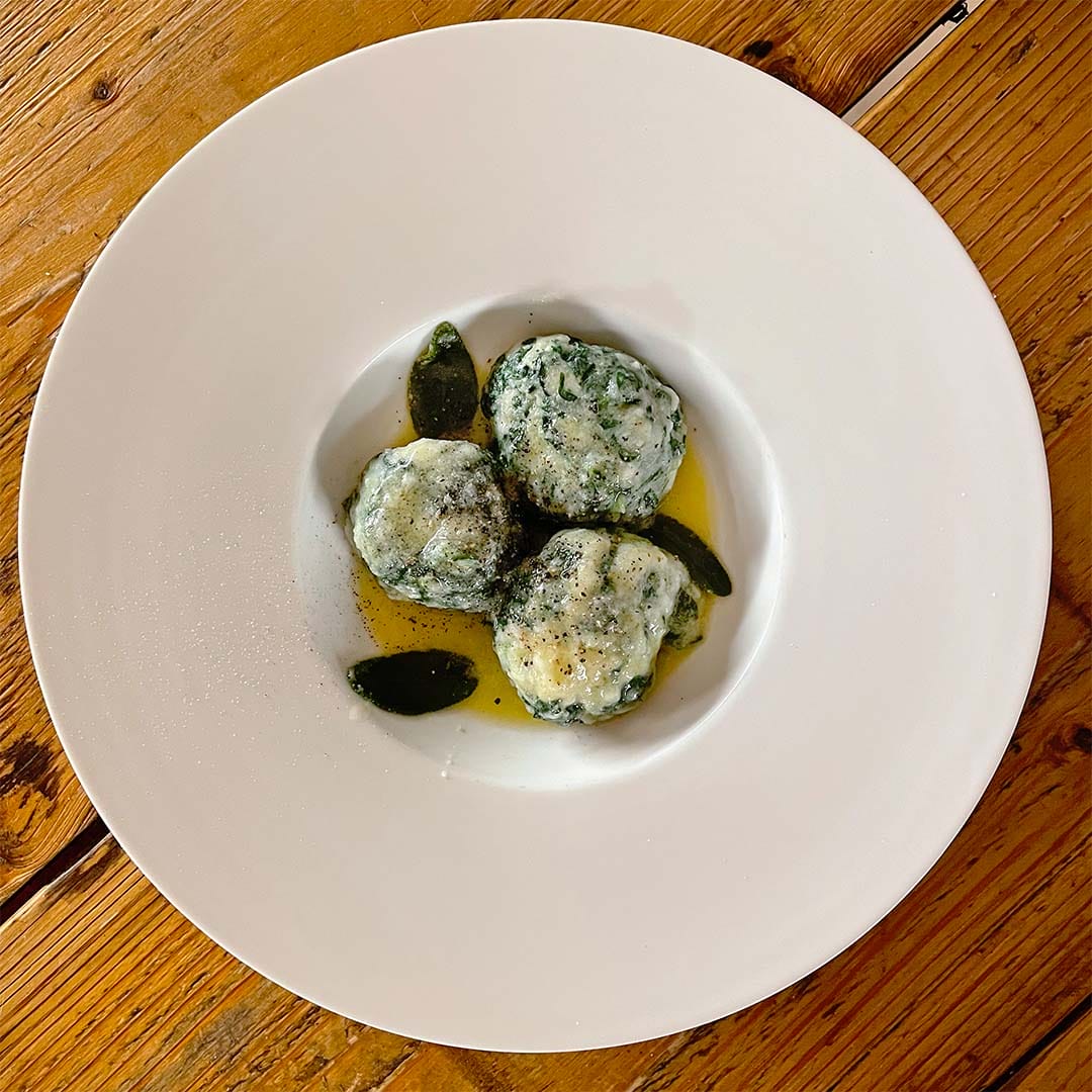 Three spinach and ricotta dumplings served on a white plate with sage leaves and melted butter, placed on a rustic wooden table.