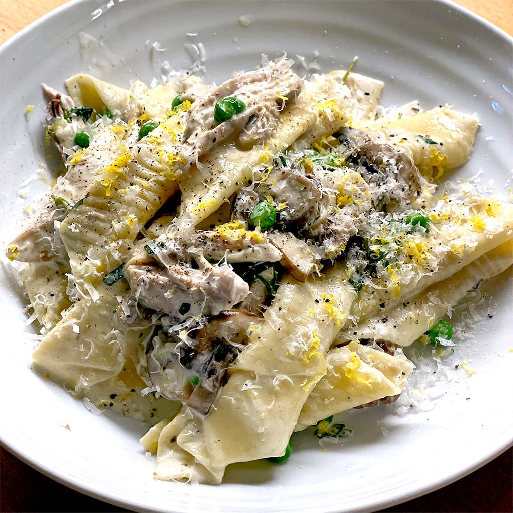 A plate of wide, homemade pasta with creamy mushroom sauce, topped with grated cheese, chopped herbs, and lemon zest, served on a white dish.