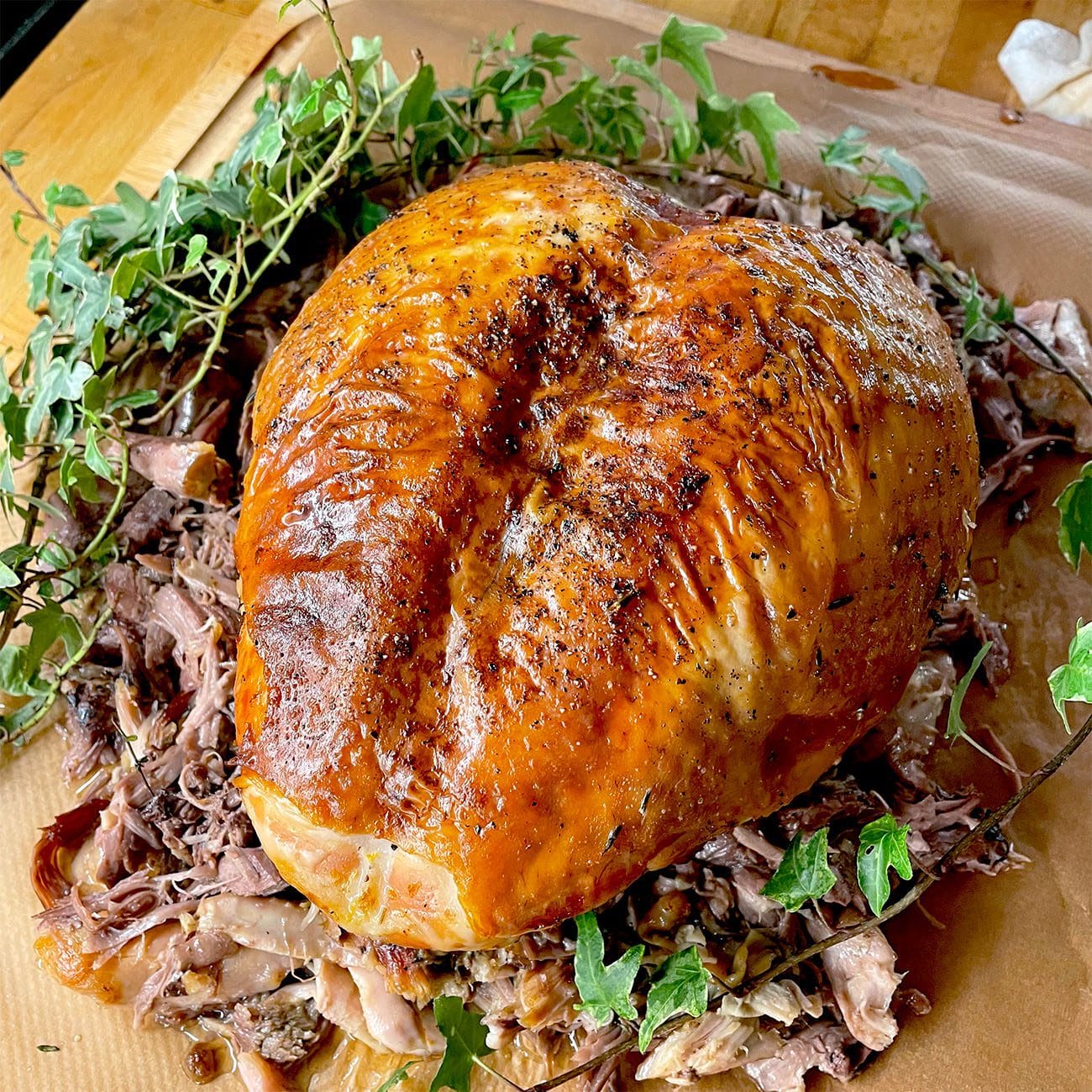 A golden-brown, roasted turkey breast is surrounded by shredded turkey meat and garnished with fresh green herbs on a wooden surface.