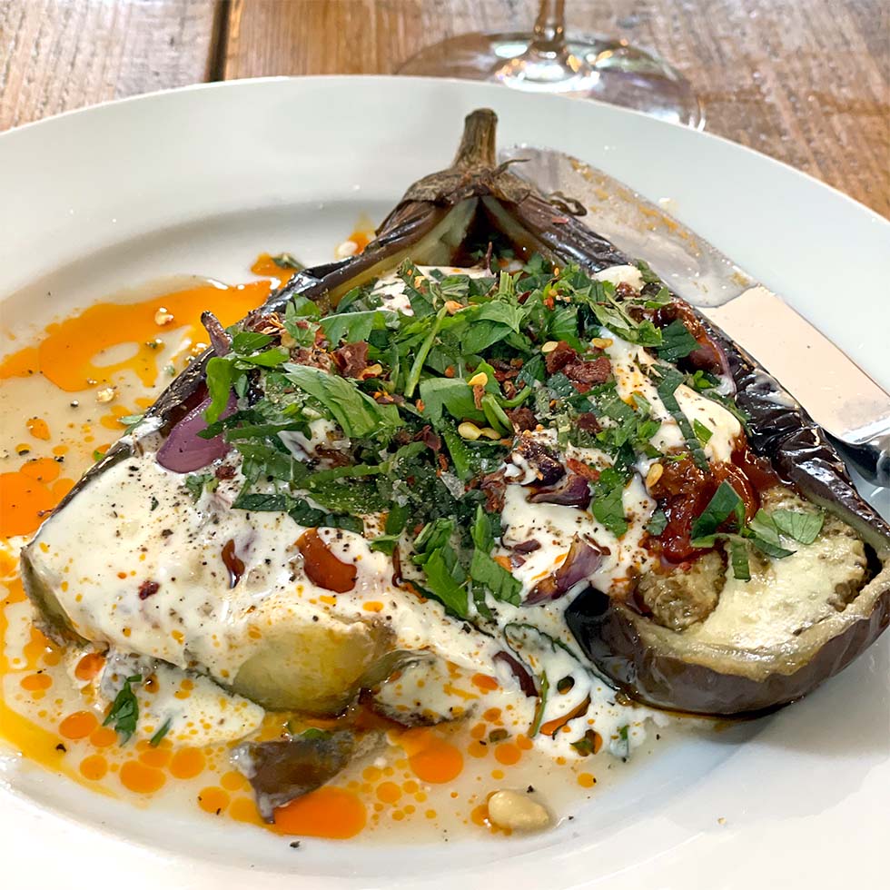 A roasted aubergine half topped with creamy yoghurt sauce, chopped fresh herbs, red pepper flakes, and a drizzle of oil, served on a white plate with a knife and fork on a wooden table.