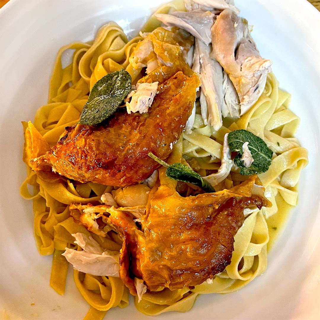 A plate of fettuccine pasta topped with roasted chicken pieces and crispy sage leaves, served on a white dish.