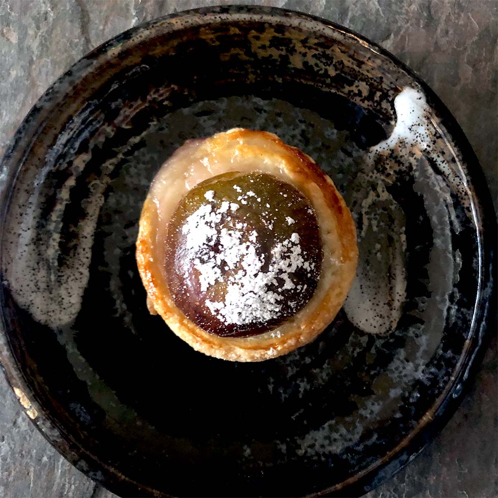 A round pastry with a glossy, dark filling dusted with icing sugar sits on a rustic, dark ceramic plate with a swirl of white sauce.