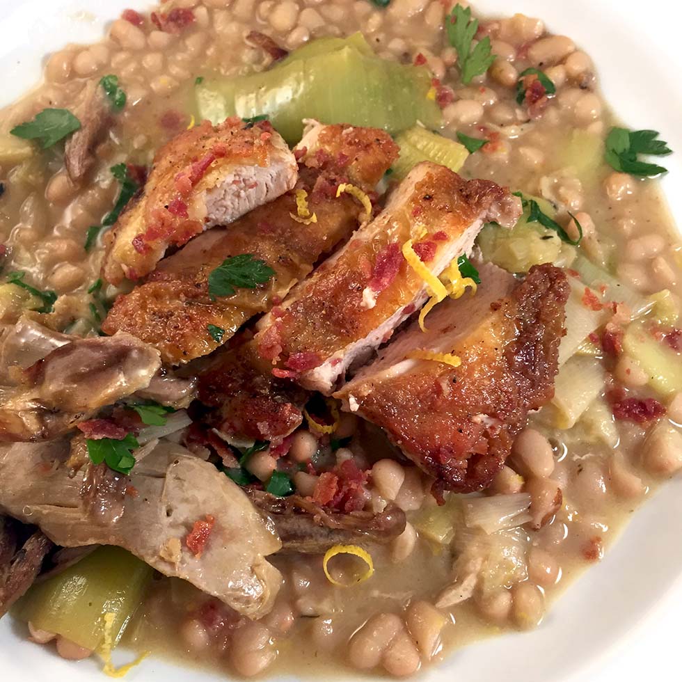 A close-up of a plate with crispy sliced chicken on top of a hearty white bean stew, garnished with fresh parsley and lemon zest. The stew contains beans, leeks, and shredded meat.