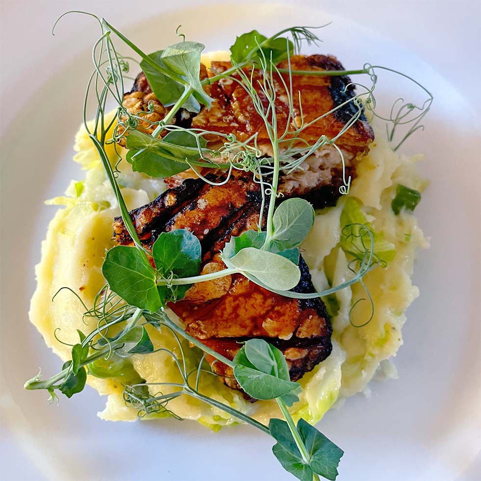 Grilled tempeh served on a bed of mashed potato, garnished with fresh pea shoots and microgreens, presented on a white plate.
