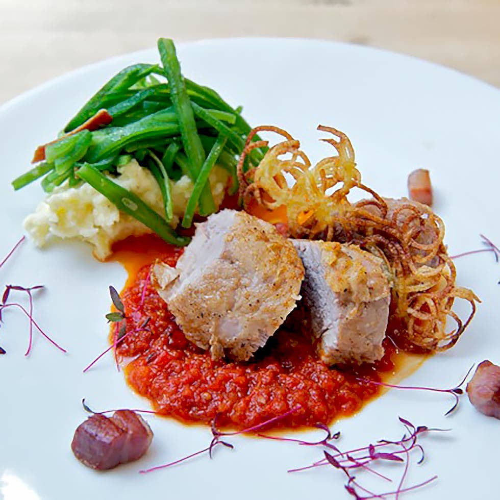 Two thick slices of cooked meat sit on a bed of red tomato sauce, served with mashed potato, green beans, crispy shoestring chips, and garnished with microgreens on a white plate.