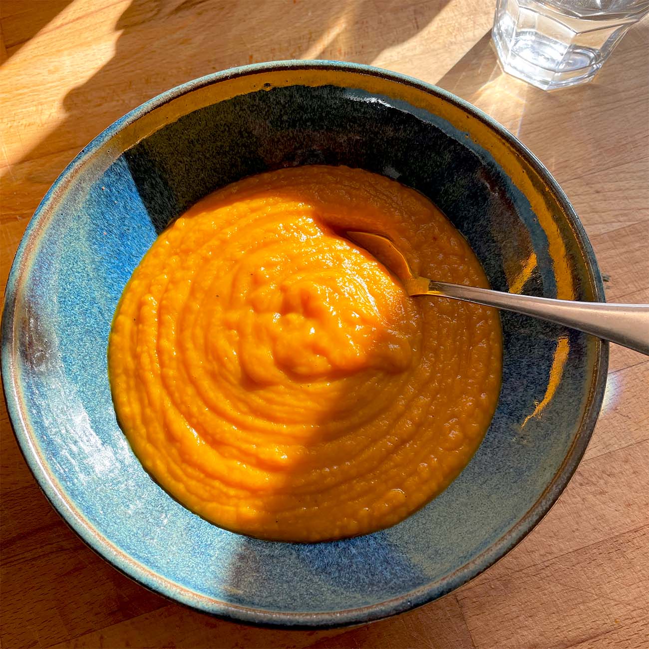 A blue ceramic bowl filled with thick, orange soup sits on a wooden table, with a spoon inside. Sunlight and shadows fall across the bowl, and a glass of water is visible in the background.