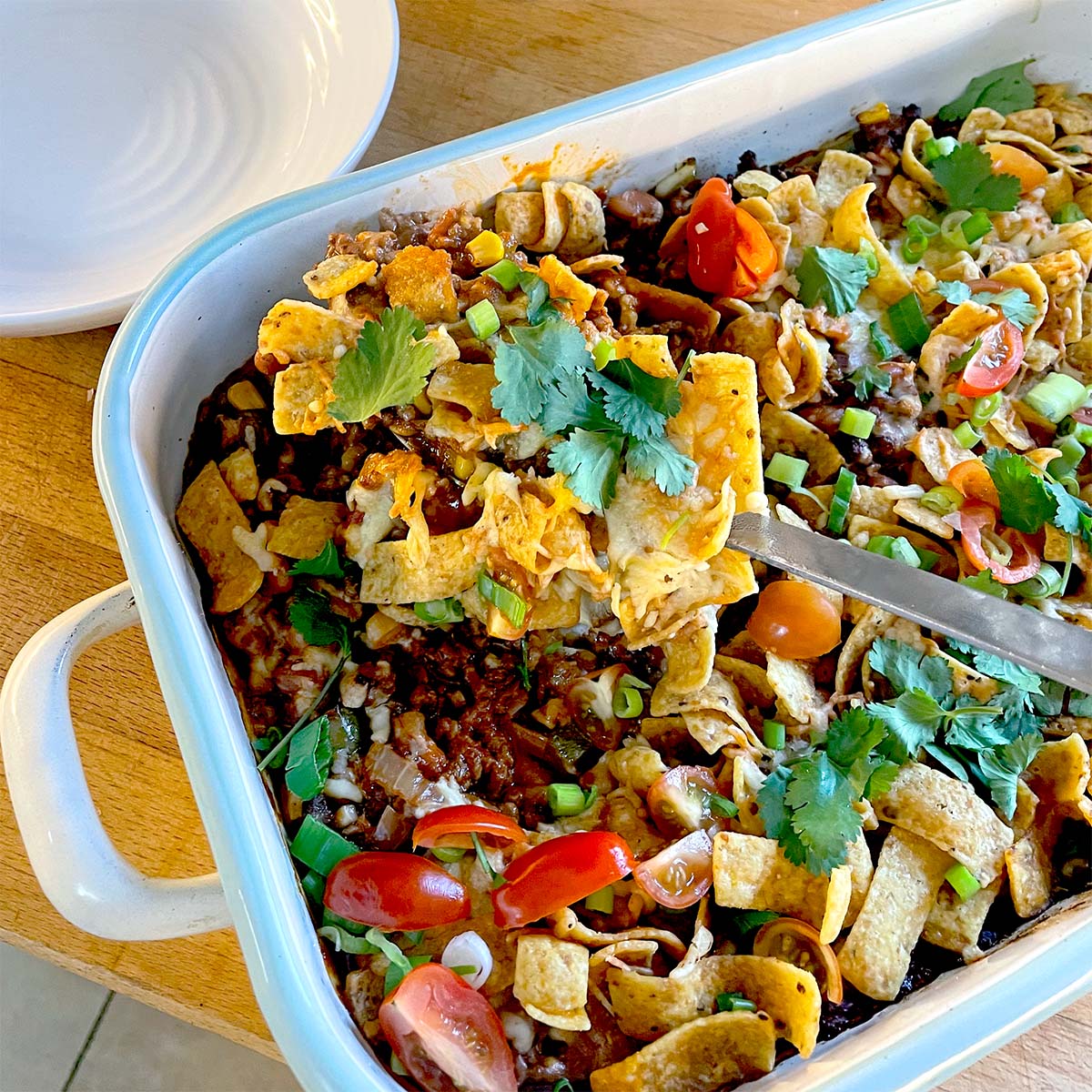 A casserole dish filled with a layered Frito pie topped with corn chips, melted cheese, minced beef, tomatoes, spring onions, and coriander. A serving is being lifted out with a spatula. A stack of white plates is nearby.