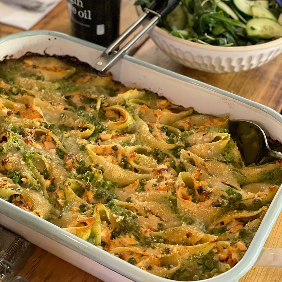 A large white baking dish filled with stuffed pasta shells topped with melted cheese and green herbs, with a serving spoon inside. In the background, there is a bowl of salad and a bottle of olive oil.