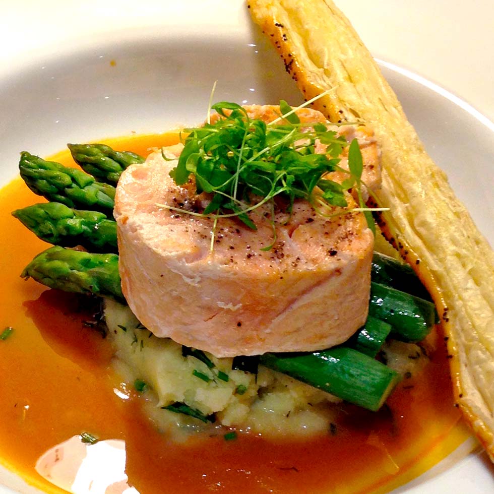 A gourmet dish featuring a round piece of salmon atop mashed potato with green asparagus spears, garnished with microgreens, served with a brown sauce and a puff pastry stick on the side.