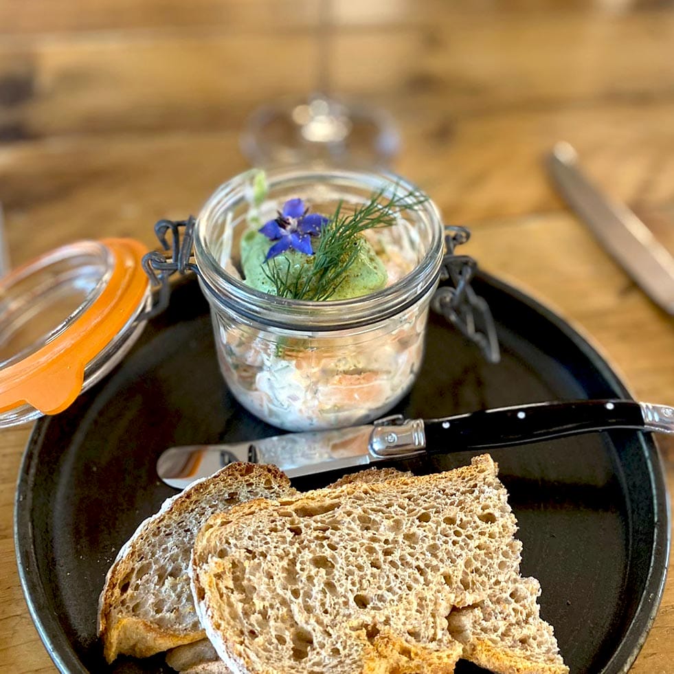 A black plate with two slices of rustic bread, a knife, and an open glass jar containing a creamy spread garnished with fresh herbs and edible flowers. The background shows a wooden table and a blurred wine glass.