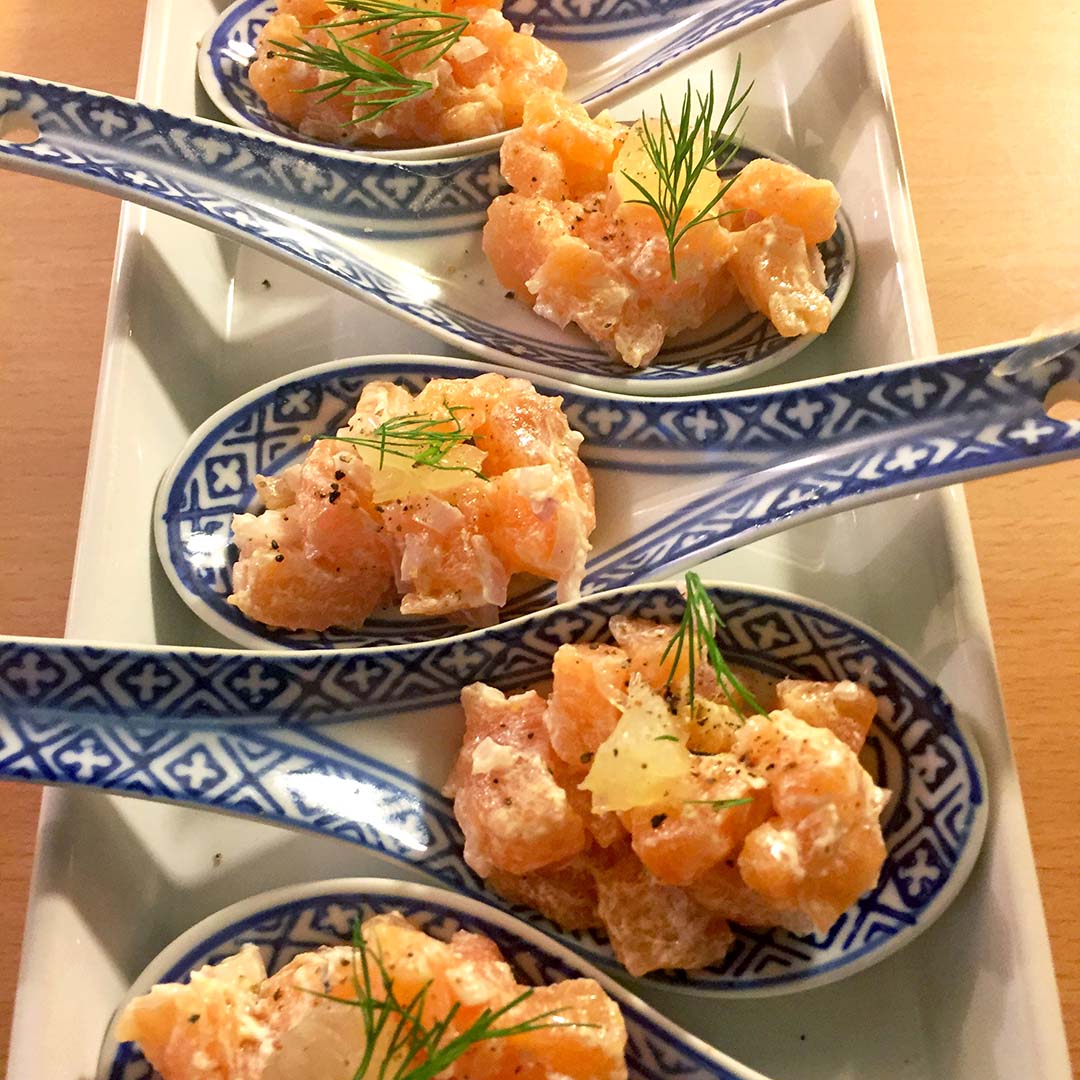 Four decorative ceramic spoons with diced raw salmon, onions, and herbs, garnished with dill and pepper, arranged neatly on a rectangular plate.