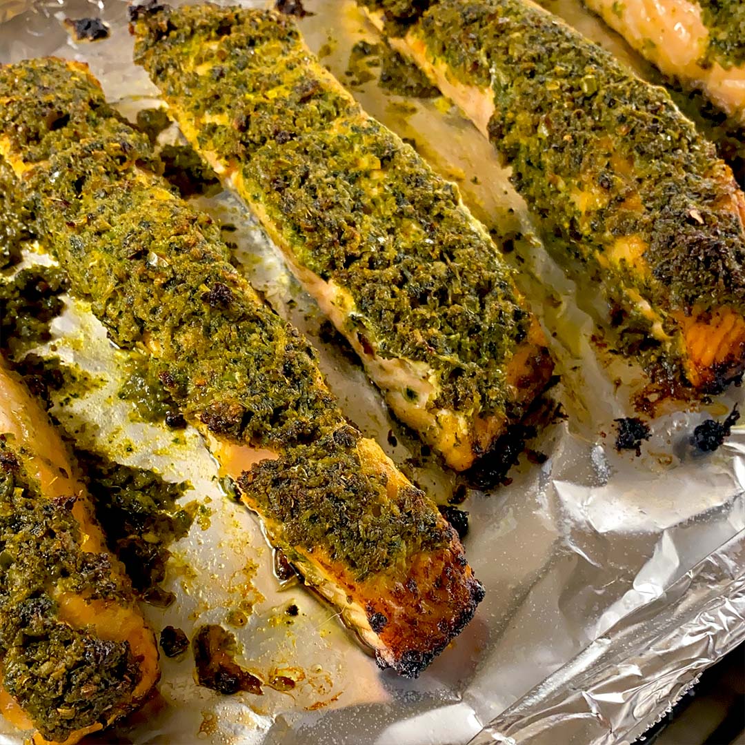 Herb-crusted salmon fillets baked on a sheet of aluminium foil, with a golden green topping and slightly crispy edges. The fish appears moist and well-seasoned.