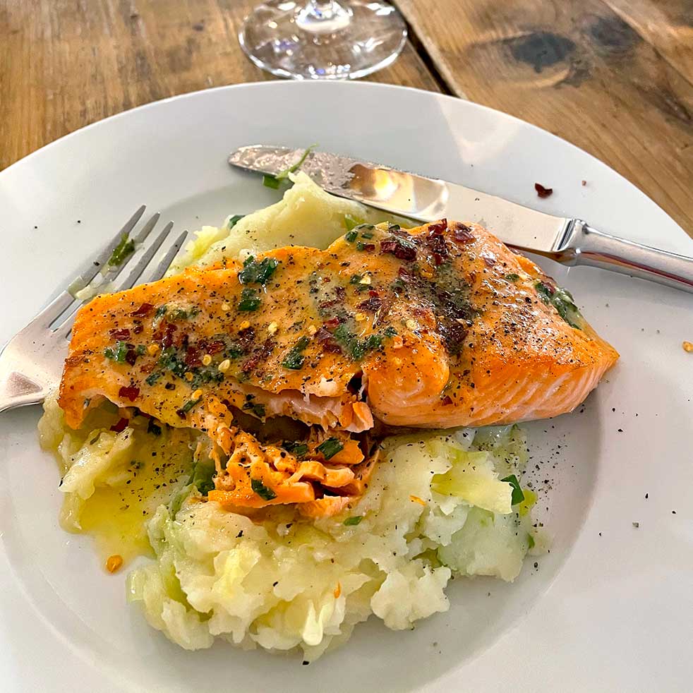 A plate of mashed potatoes topped with a seasoned, baked salmon fillet, garnished with herbs and spices. A fork and knife rest on the plate, and a glass is visible in the background on a wooden table.