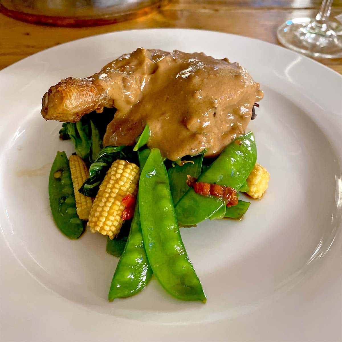 A roasted chicken leg topped with creamy brown sauce, served on a bed of sautéed mangetout, baby sweetcorn, and greens on a white plate.