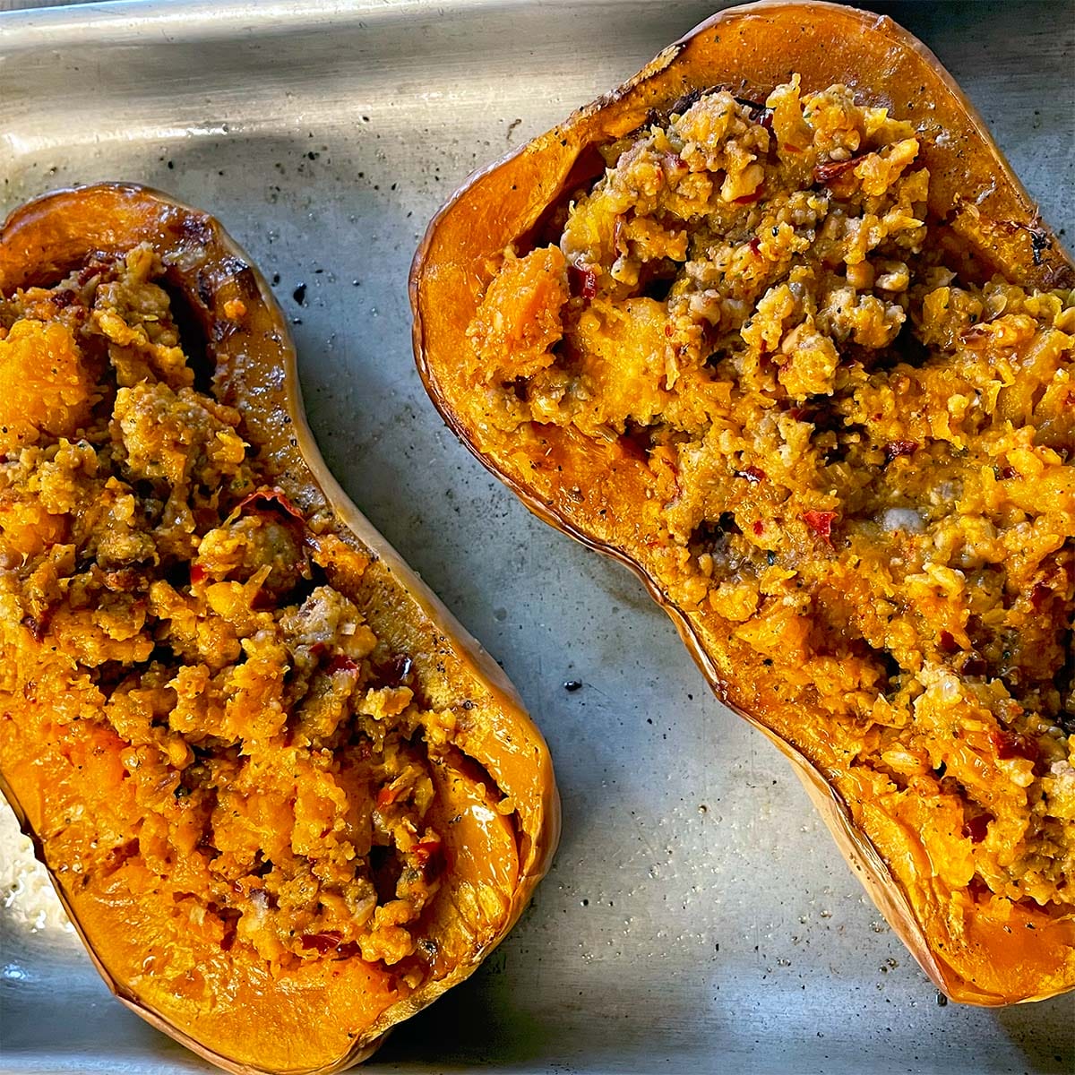 Two halves of butternut squash are filled with a crumbly, golden brown stuffing and baked on a metal tray. The stuffing appears to include grains and vegetables, giving a textured appearance.